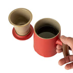 Hand holding red porcelain mug with wooden handle next to beige infuser and matching lid on white background