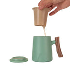 Hand pouring tea from beige infuser into green porcelain mug with wooden handle and matching lid on white background