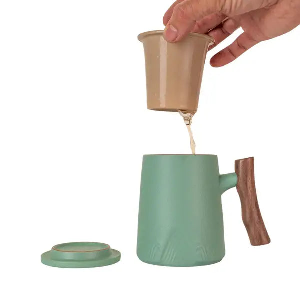 Hand pouring tea from beige infuser into green porcelain mug with wooden handle and matching lid on white background