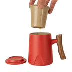Red porcelain mug with wooden handle, beige infuser dripping tea, and matching lid on white background