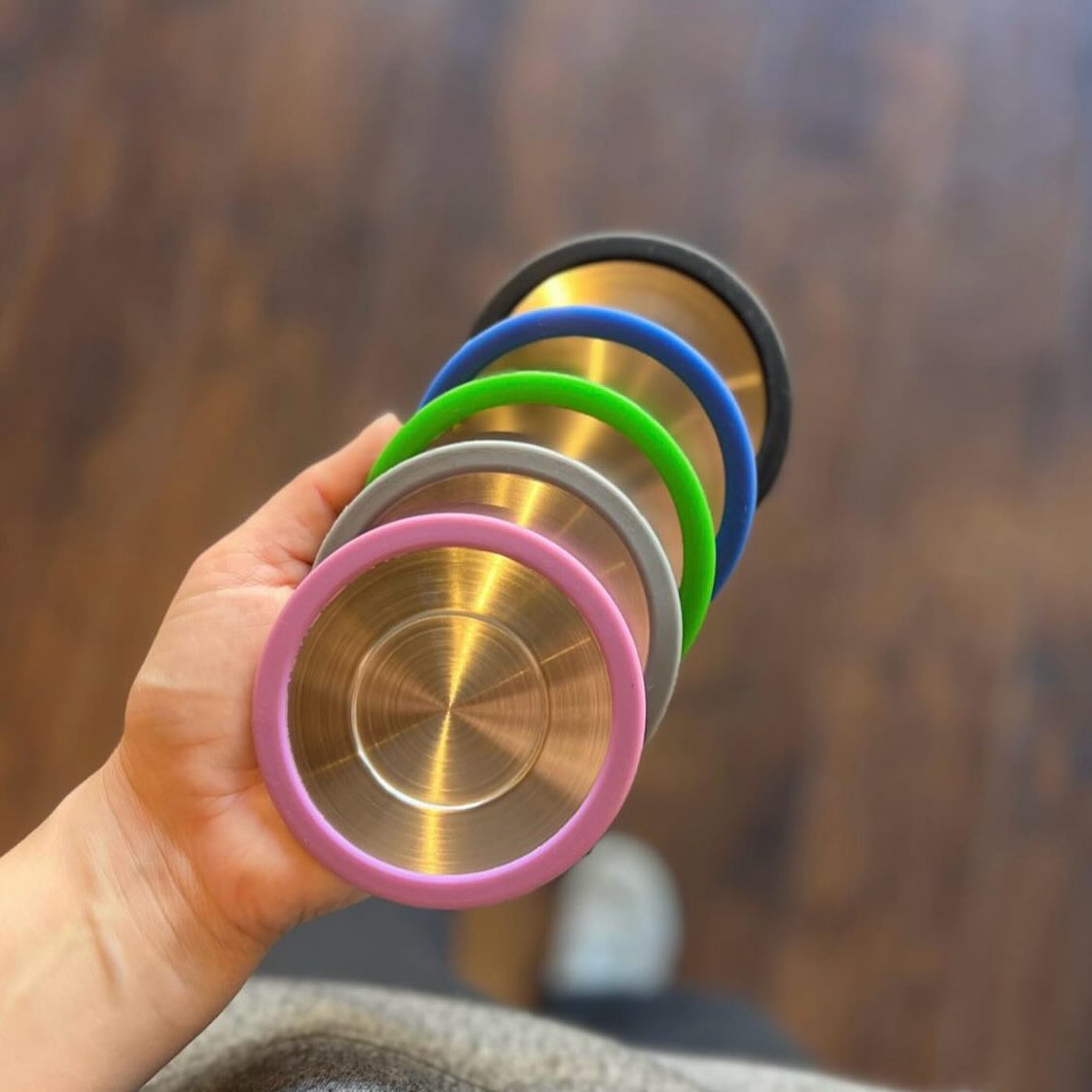 Close-up of stainless steel tea basket infusers with colorful silicone rims held in hand against wooden floor background