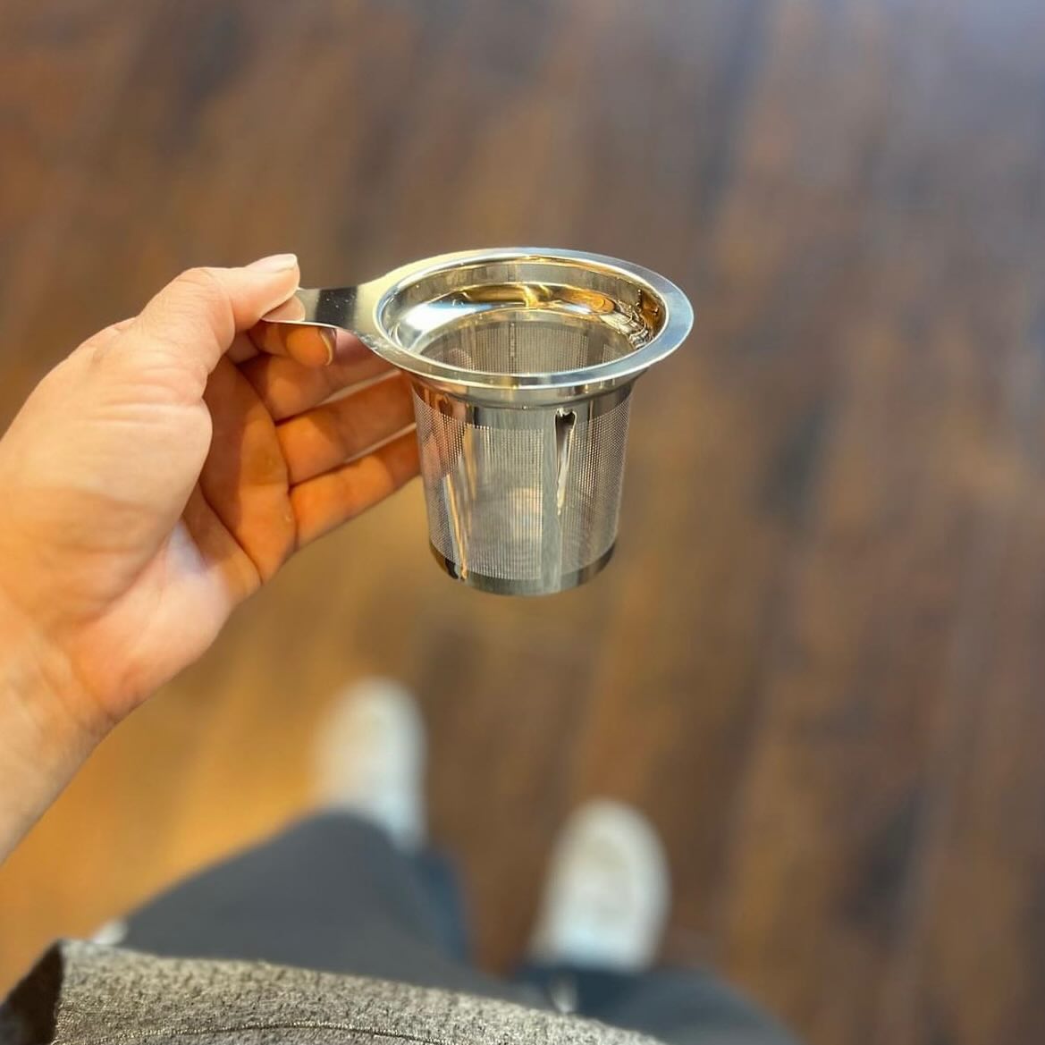 Hand holding stainless steel tea basket infuser with fine mesh and handle over wooden floor background