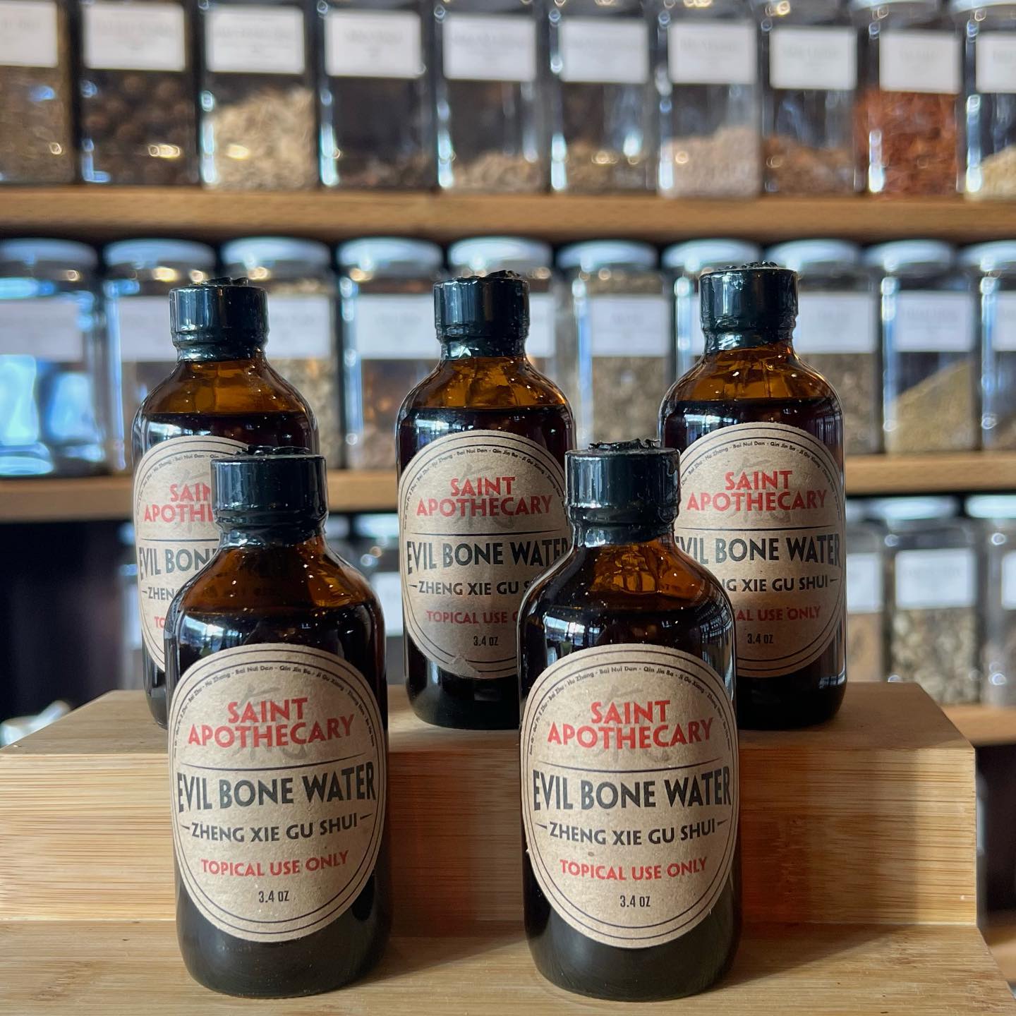 Five amber bottles of Evil Bone Water topical liniment with vintage labels on wooden surface and herbs jars background