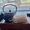 Cloth pouch of loose leaf tea bags resting on a wooden tray with a textured black cast iron teapot in the background.