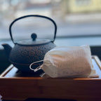 Cloth pouch of loose leaf tea bags resting on a wooden tray with a textured black cast iron teapot in the background.