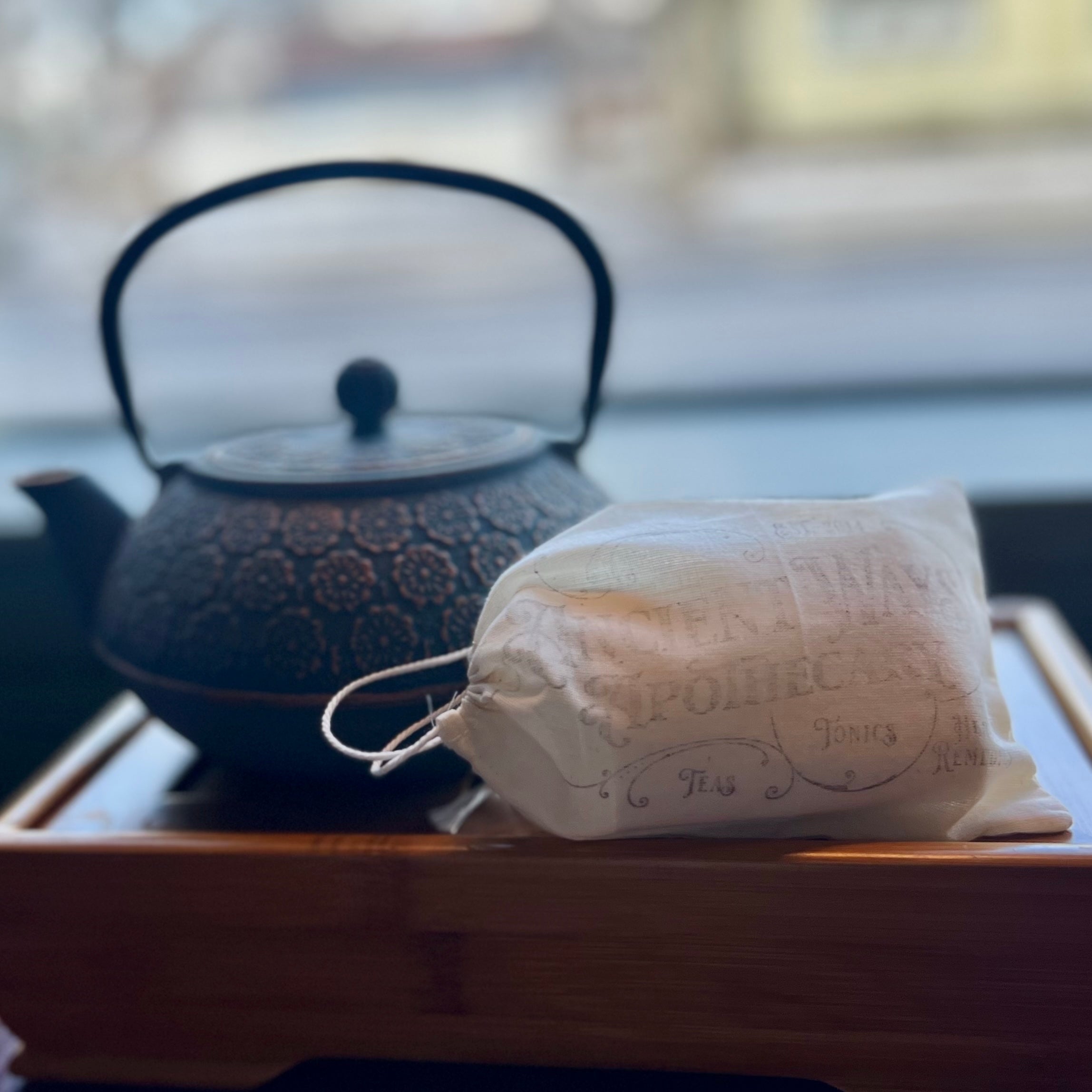 Cloth pouch of loose leaf tea bags resting on a wooden tray with a textured black cast iron teapot in the background.