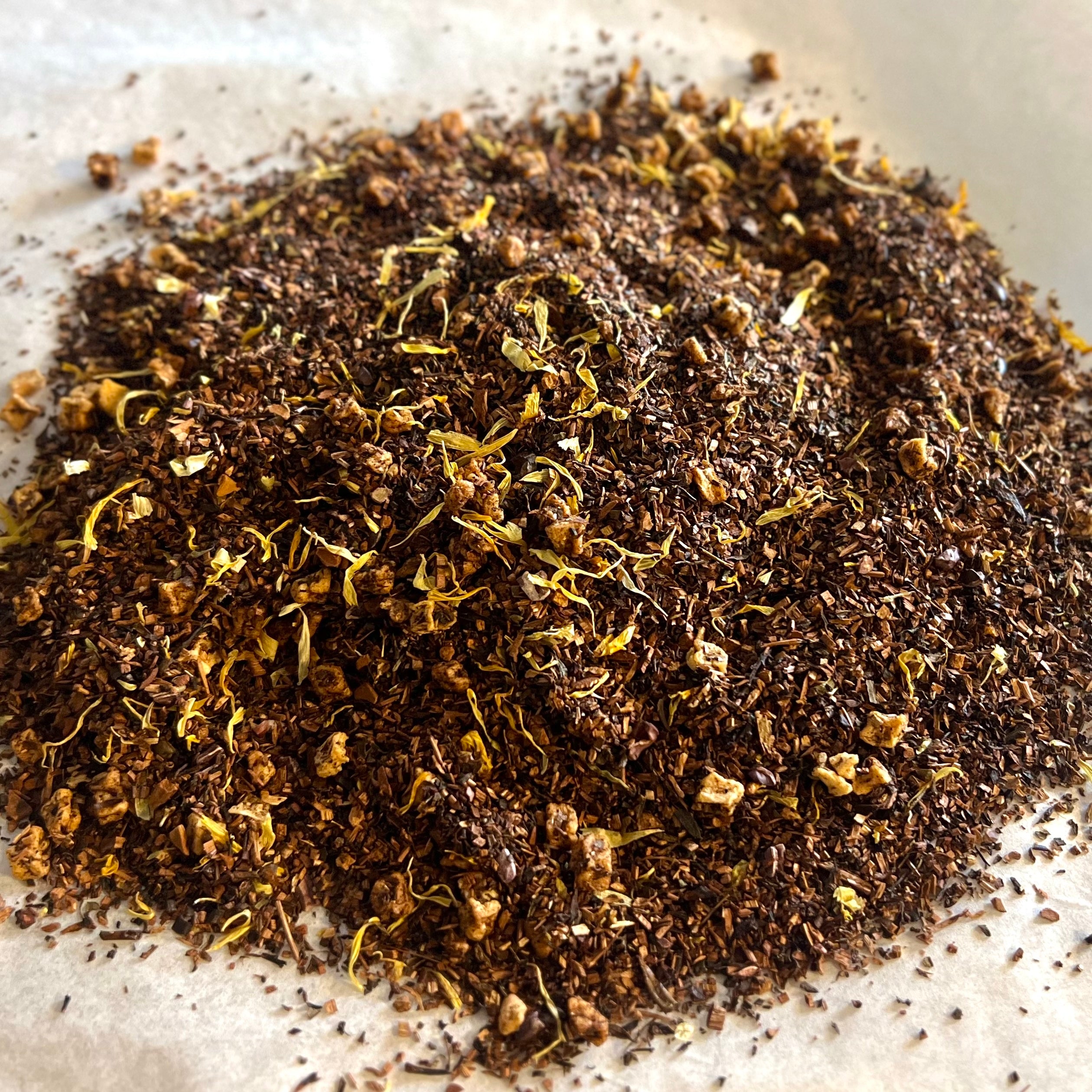 Close-up of loose Banana Bread tea blend with honeybush and dried flower petals on a light parchment surface