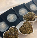 Four black tea pouches laid out with heart-shaped bowls of loose herbal tea blends featuring dried leaves and petals