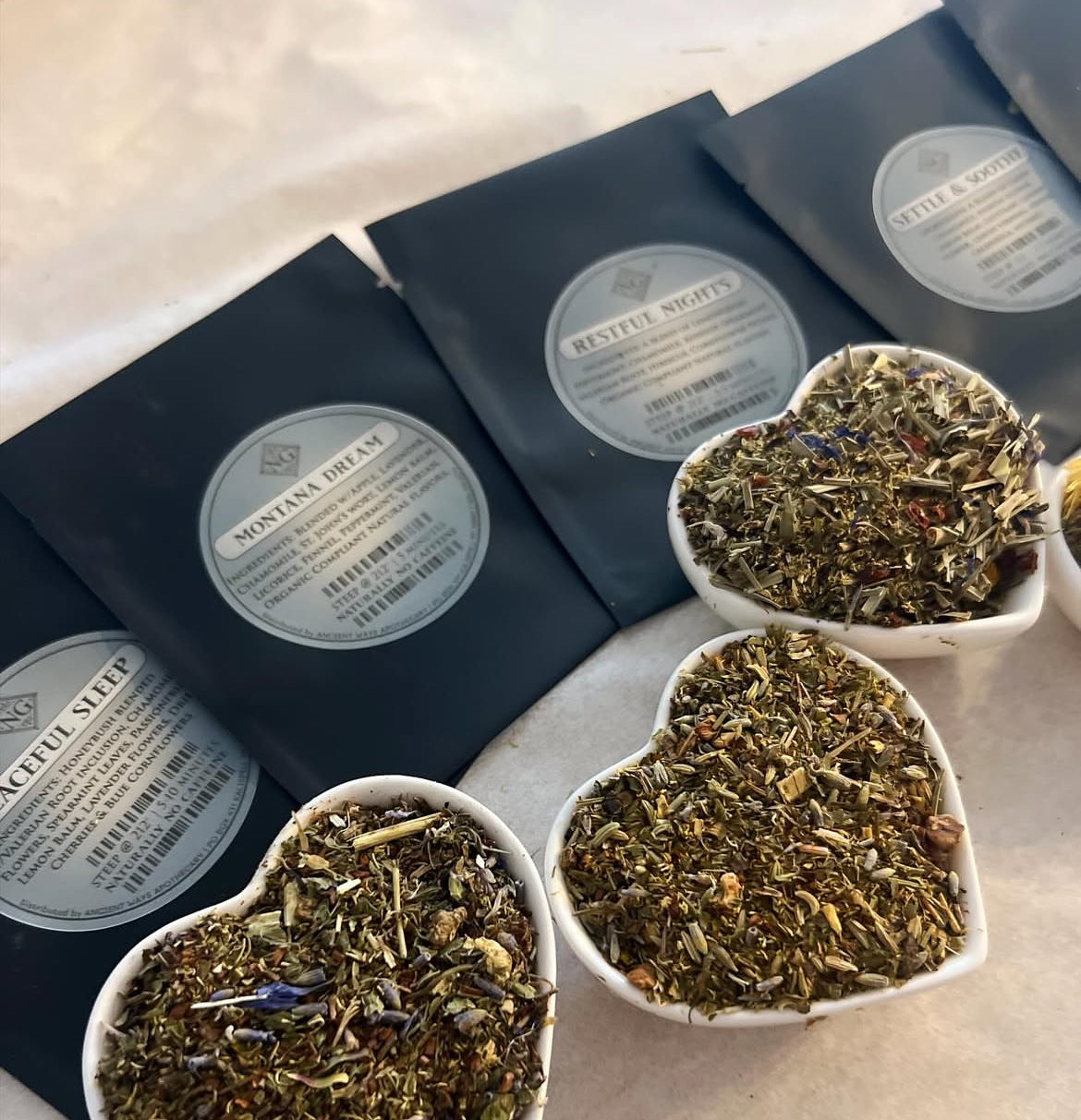 Four black tea pouches laid out with heart-shaped bowls of loose herbal tea blends featuring dried leaves and petals