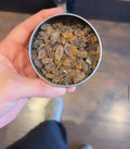 Close-up of amber-colored tea sugar crystals in a round metal container held by a hand over wooden floor background