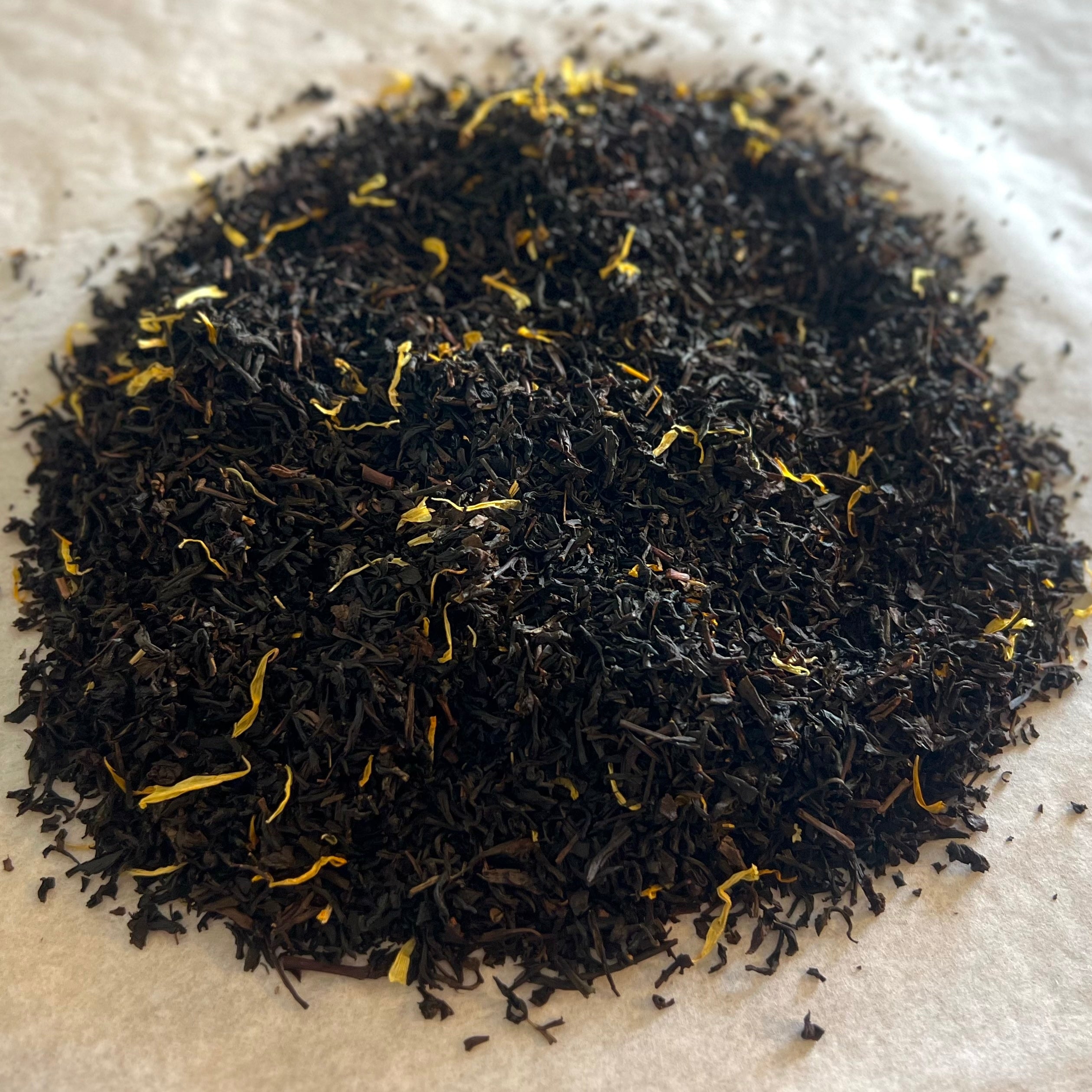 Close-up of loose Creme Brûlée black tea blend with dark leaves and delicate yellow flower petals on white surface.