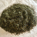 Close-up of loose Jade Court green tea leaves with fine, dark green Sencha bits on a light parchment surface.