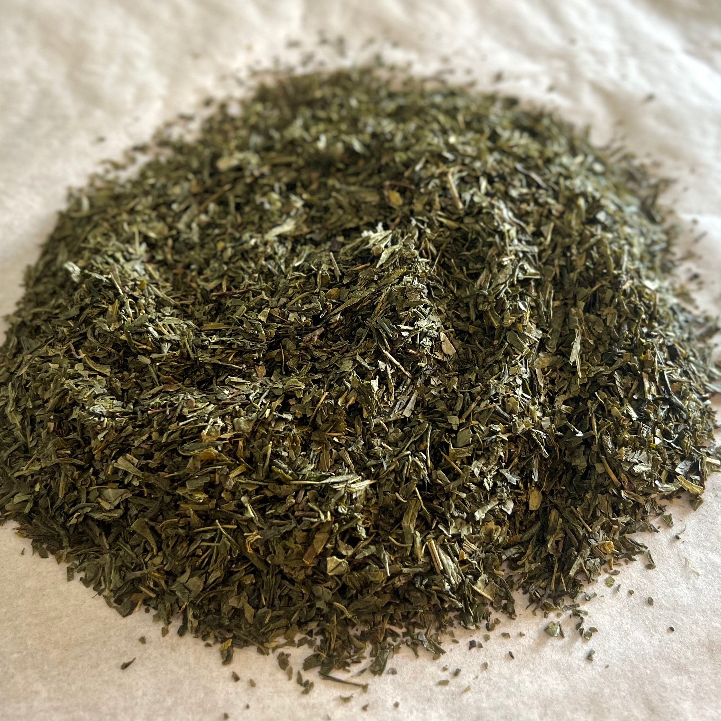 Close-up of loose Jade Court green tea leaves with fine, dark green Sencha bits on a light parchment surface.
