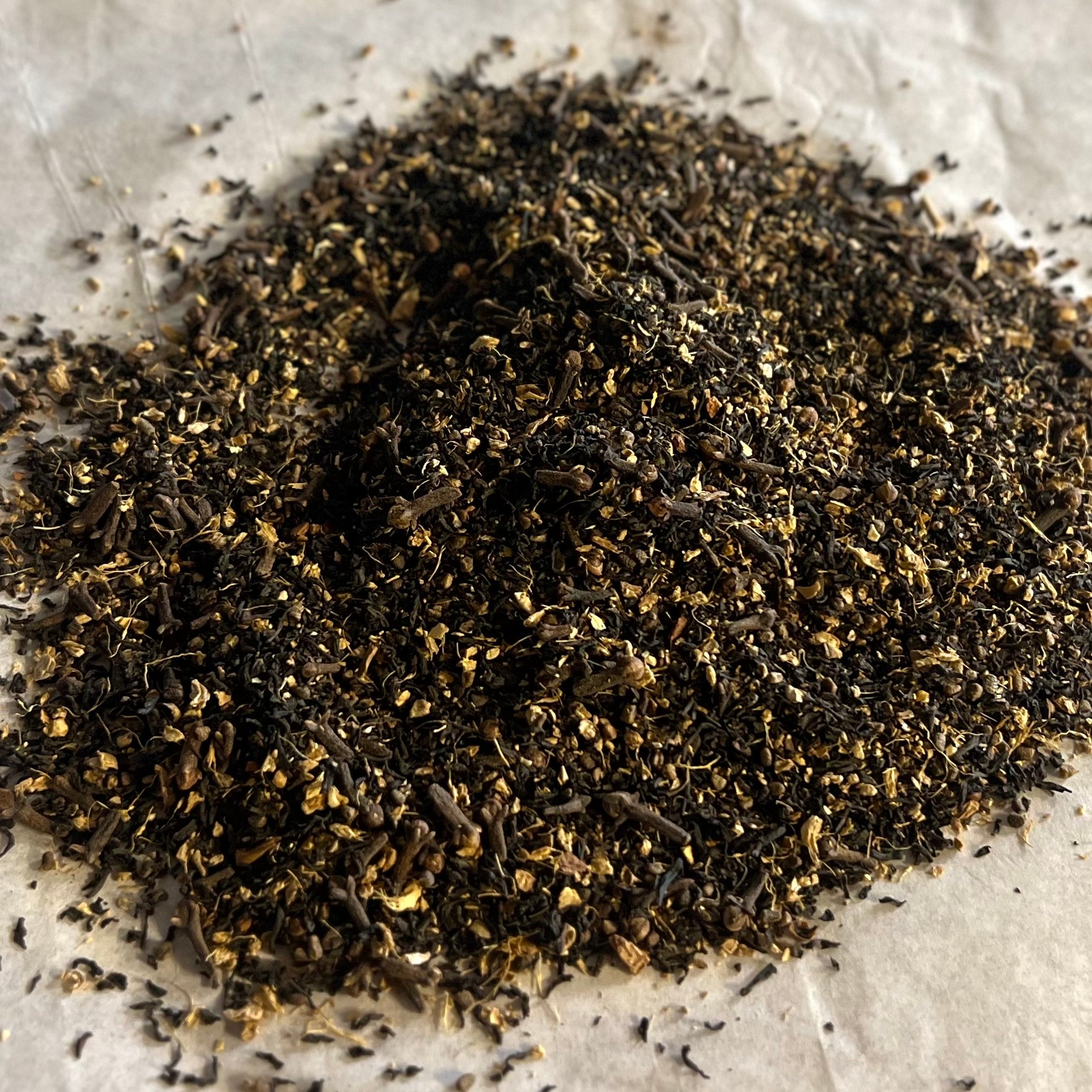 Close-up of loose chai blend with black tea leaves, spices, and dried ginger pieces on parchment background