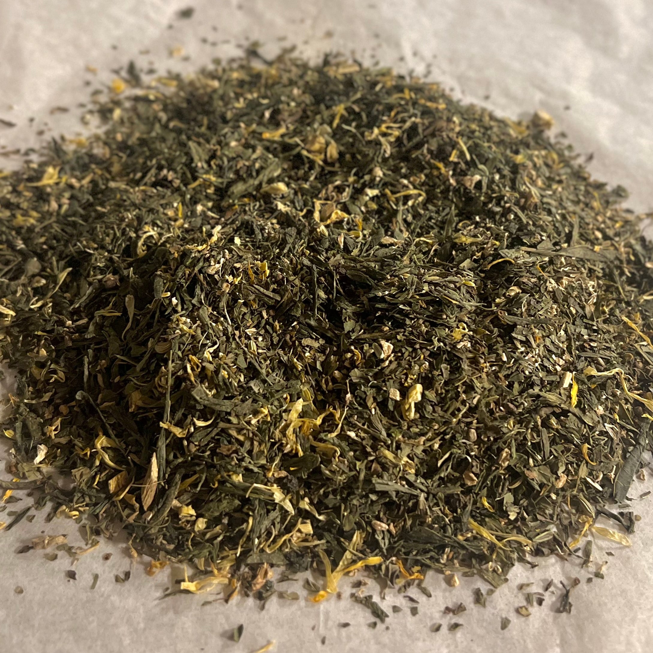 Close-up of Recover Restore loose green tea blend with mixed herbs and yellow flower petals on parchment paper.