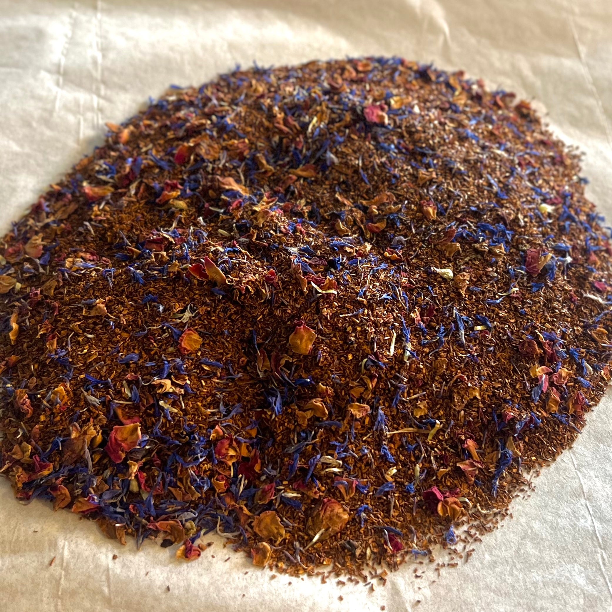 Close-up of Relax & Joy rooibos tea blend with caramel, almond, vanilla, rose petals, and blue cornflower petals on parchment paper.