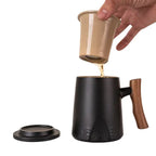 Black porcelain mug with wooden handle and ceramic infuser pouring tea, alongside a matching black lid on white background
