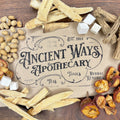 Overhead shot of dried herbs, roots, sugar cubes, and fruit surrounding vintage style Ancient Ways Apothecary label on wood table.