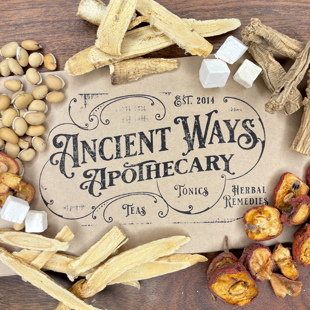 Overhead shot of dried herbs, roots, sugar cubes, and fruit surrounding vintage style Ancient Ways Apothecary label on wood table.
