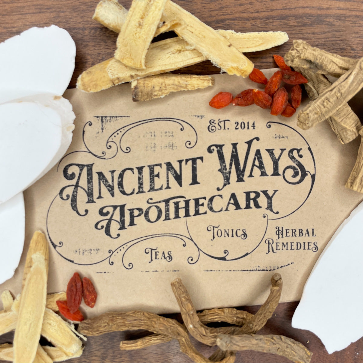 Dried herbal ingredients including Huang Qi, Dang Gui, Goji berries, and Shan Yao surrounding Ancient Ways Apothecary label on wood surface