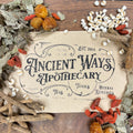 Vintage style paper label reading Ancient Ways Apothecary surrounded by dried herbs and roots on wooden surface