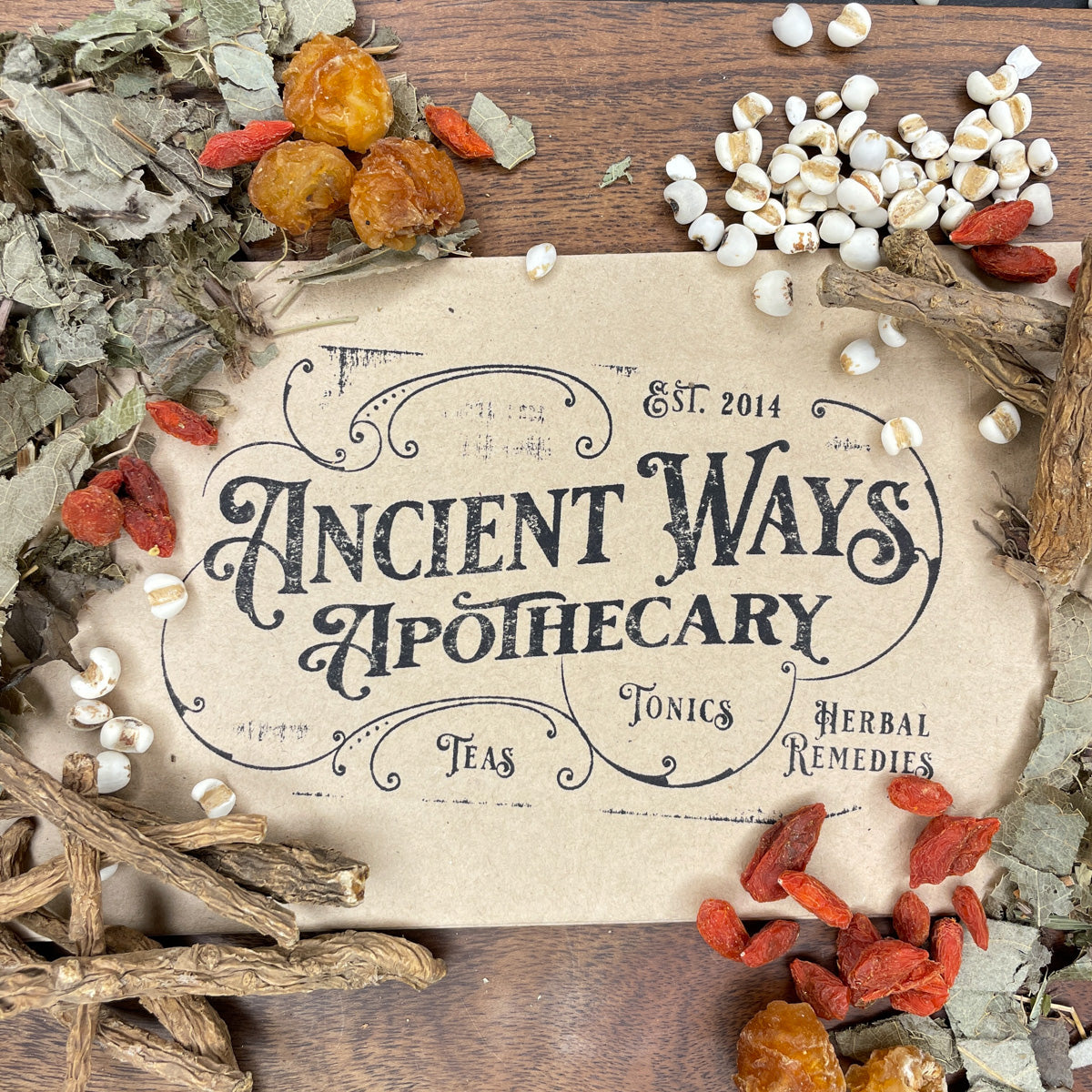 Vintage style paper label reading Ancient Ways Apothecary surrounded by dried herbs and roots on wooden surface