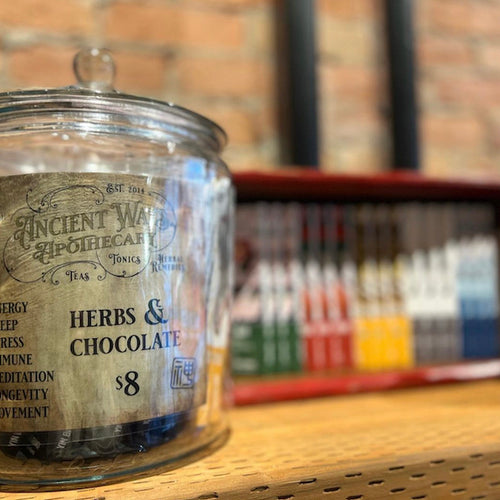 Glass apothecary jar labeled "Herbs & Chocolate" with blurred colorful packaged bars in the background on wood shelf
