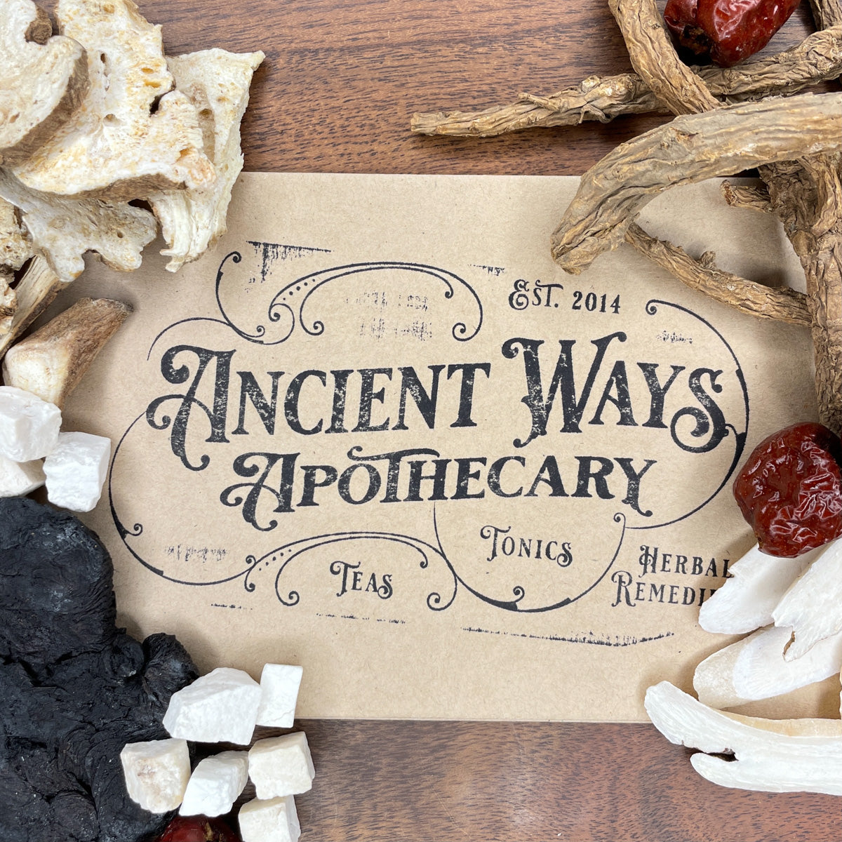 Vintage-style Ancient Ways Apothecary label surrounded by dried Chinese herbs including Dang Gui and Hong Zao on wood surface