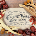 Dried traditional herbs including Huang Qi, Bai Zhu, Goji berries, and reishi mushrooms around an Ancient Ways Apothecary card on wood